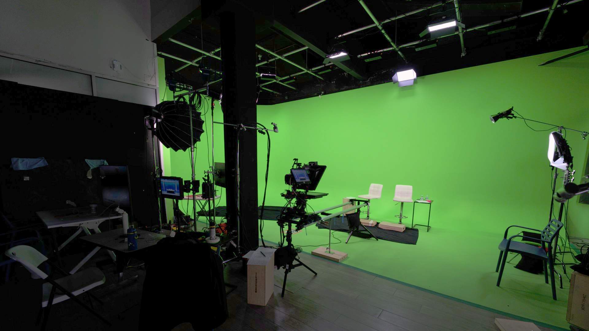 Picture of a green screen stage with lights, screens, a camera, and chairs